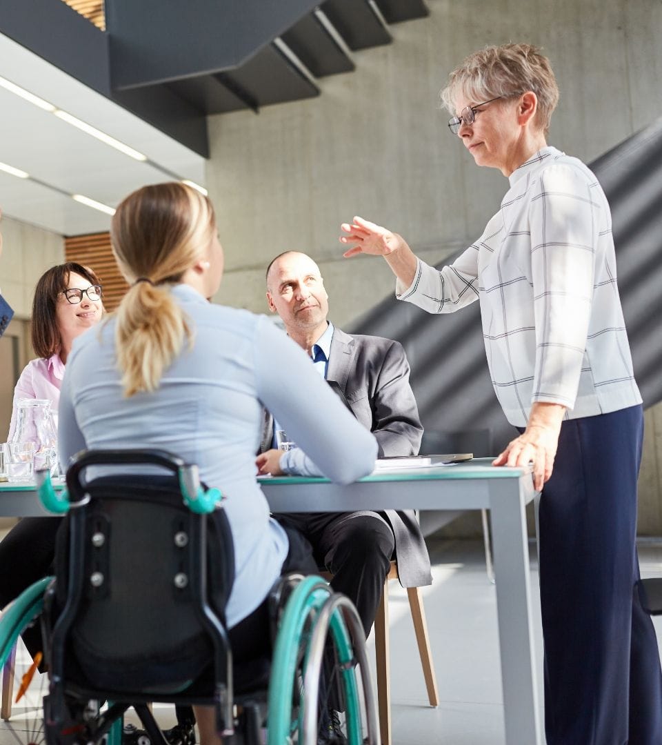 1 standing woman talking to 3 persons sitting on a table