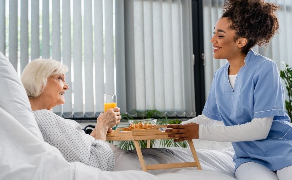 old person lying in bed getting served food from a friendly looking women