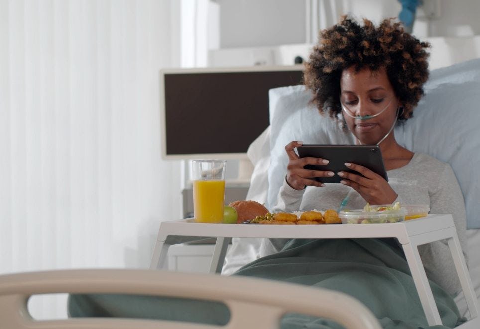 woman lying in hospital bed looking at a tablet with foodservings in front of her