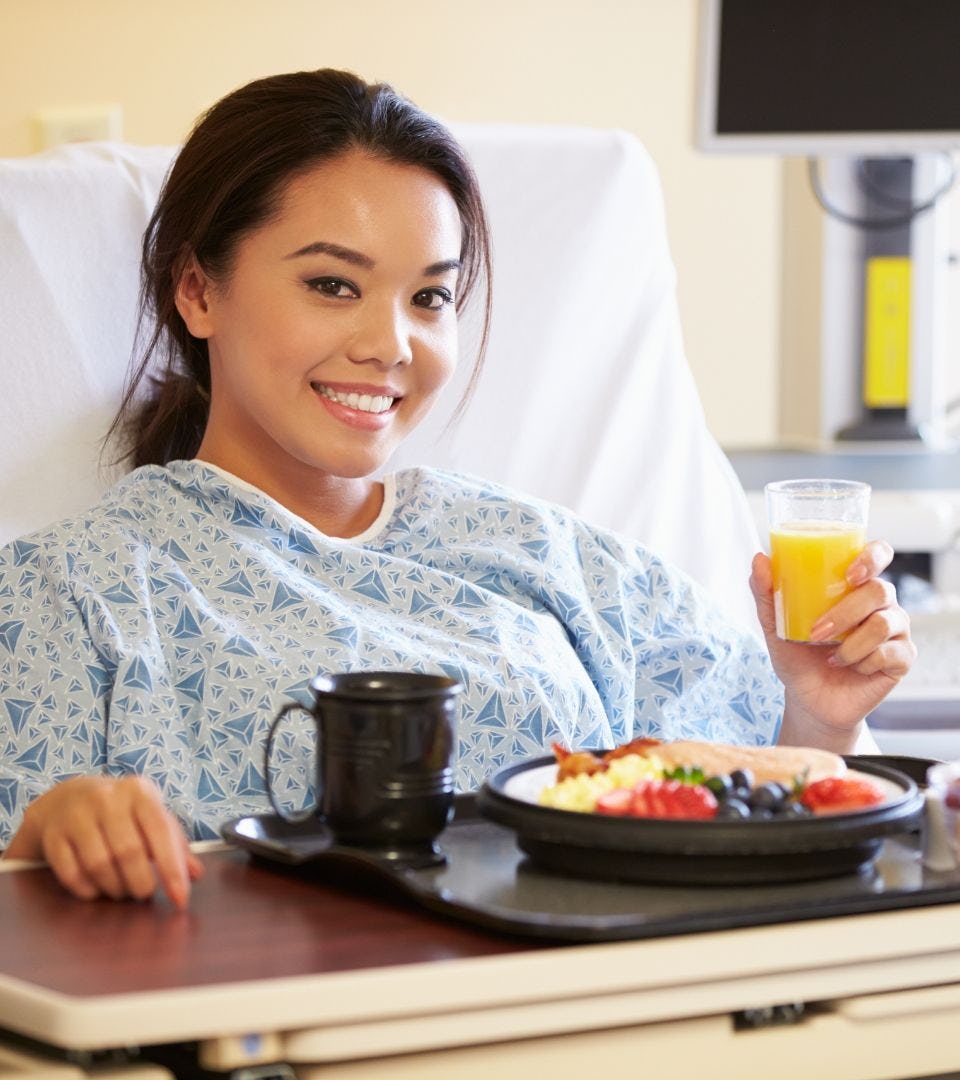 A asian women lying in a hospital the bed with a table of food on top of it